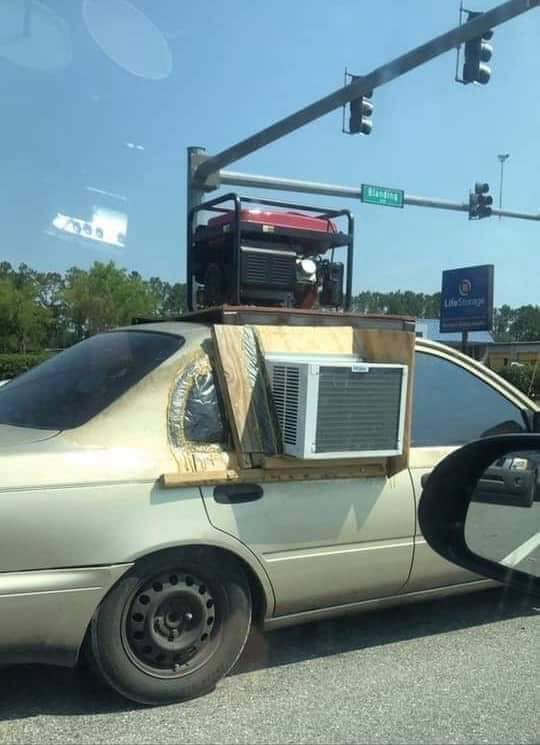 This meme is an image of a car with a makeshift air conditioning unit installed on the back window. The unit is a small window air conditioner mounted on a piece of plywood, held in place by a strap. The text "I'm not lazy, I'm just conserving energy" is written on a piece of paper stuck to the window.